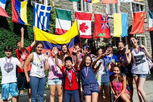 A group of children stand together, beaming with smiles as they enthusiastically wave at the camera. Behind them, a colorful display of flags from various countries around the world flutters in the breeze.