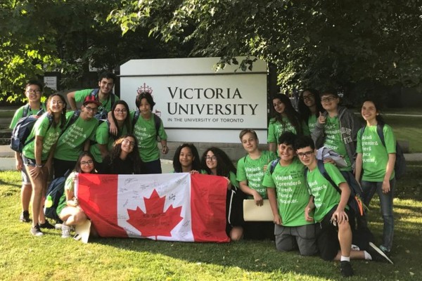 A group of teenagers, all wearing matching green t-shirts, proudly hold up a Canadian flag. They stand in front of the Victoria University sign, with bright smiles on their faces.