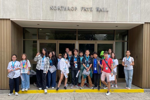 A cheerful group of teenagers stands proudly in front of the main entrance to Northrop Frye Hall on the Victoria University campus.