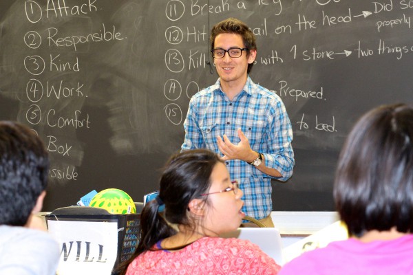 A young instructor stands confidently in front of a chalkboard, engaging a group of attentive teenagers. 