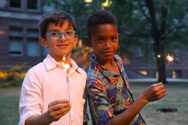 Two young boys stand on the Victoria University campus quad in the evening, holding sparklers that light up the darkening sky. 