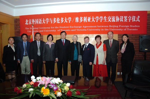 An elderly Isabel Crook stands proudly alongside a group of individuals in front of a striking red banner. The banner prominently displays the text: 'The Signing Ceremony for the Student Exchange Agreement between Beijing Foreign Studies University and University of Toronto and Victoria University in the University of Toronto.' The group appears engaged and united, representing a significant moment in international academic collaboration.